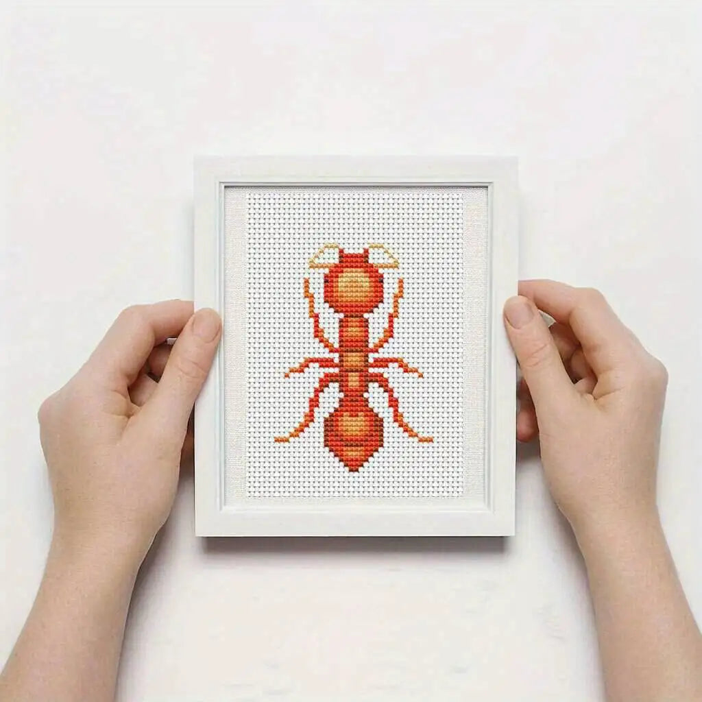 Framed cross-stitched ant pattern held by hands on a white background